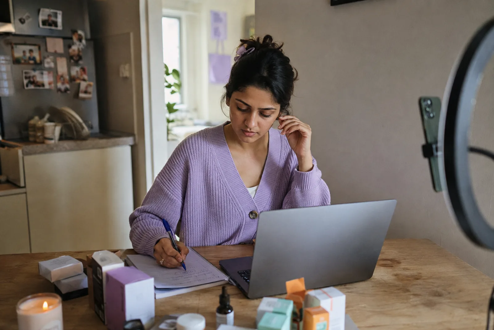 A creator at her kitchen table planning brand outreach: laptop open, a handwritten shortlist, small product boxes, and a ring light beside her.