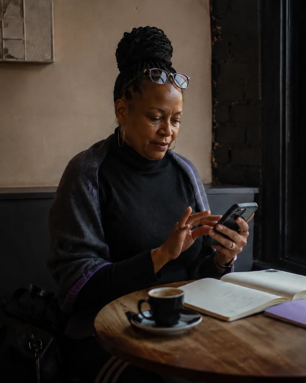 A creator at a café window table, reading glasses pushed up, scrolling her phone with an espresso and an open notebook beside her.