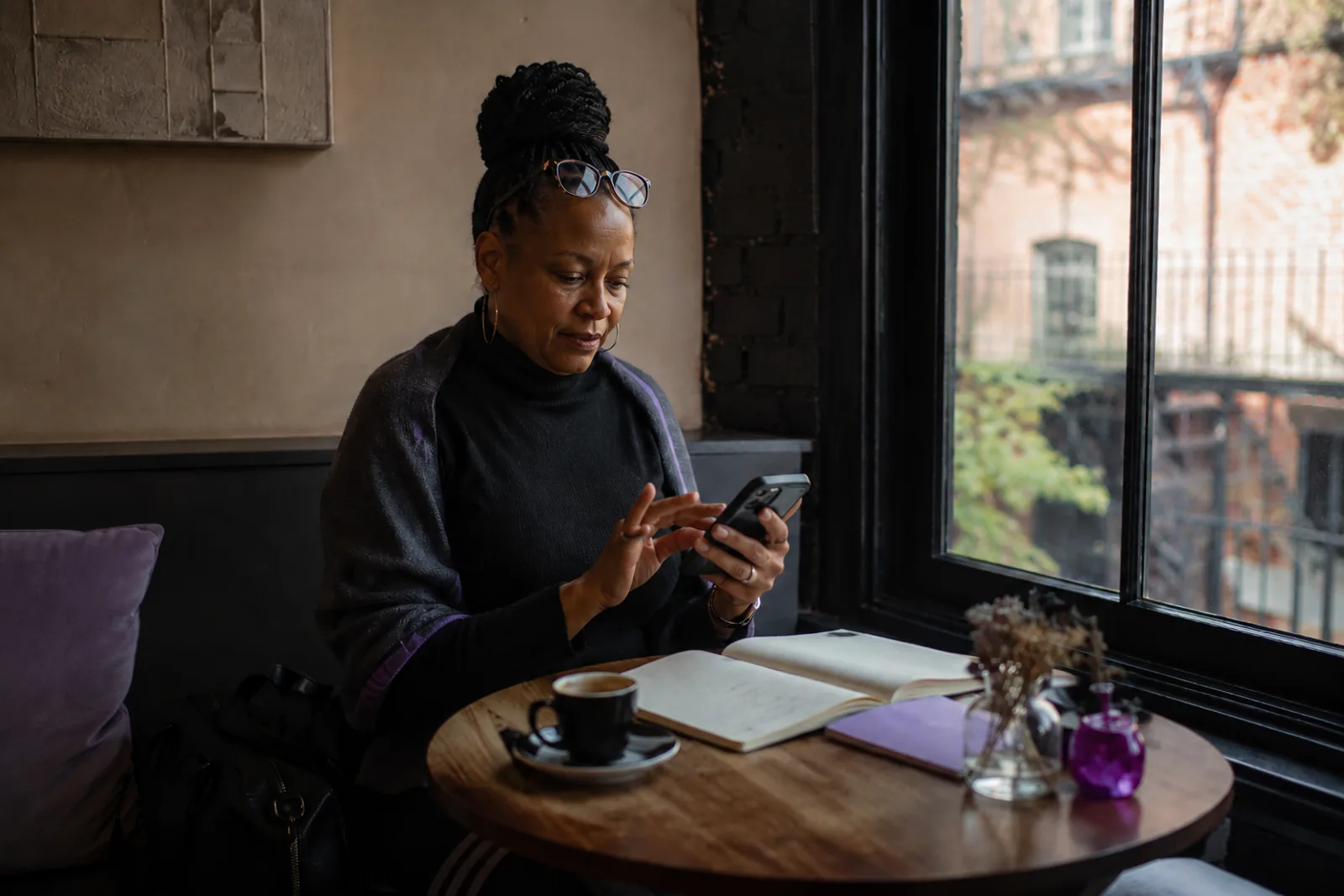 A creator at a café window table, reading glasses pushed up, scrolling her phone with an espresso and an open notebook beside her.
