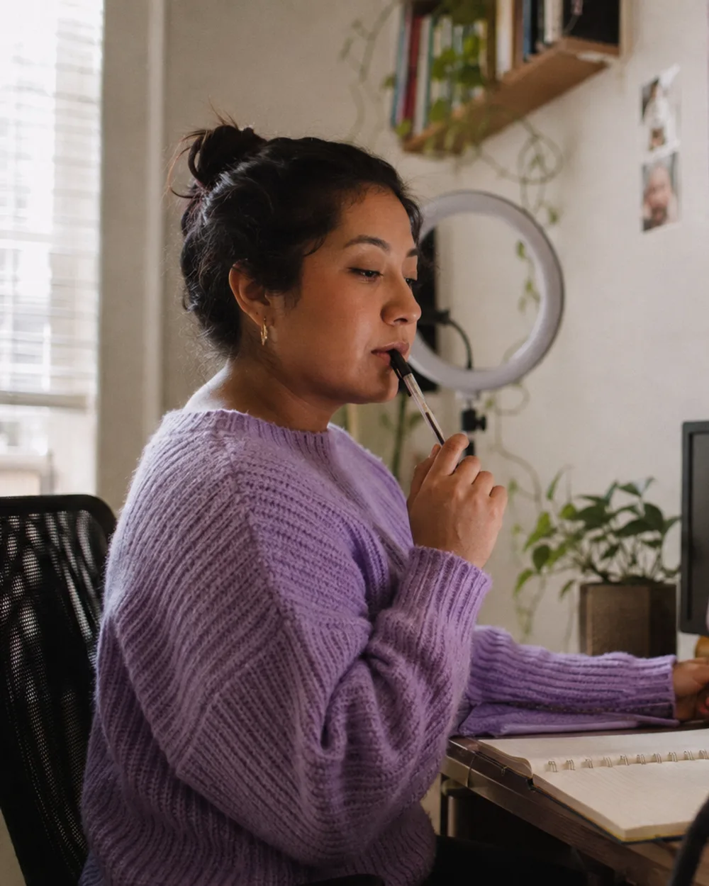 A creator at her desk with a pen at her lips, a blank checklist taped to the wall, two monitors and a phone on a tripod, thinking through the next deliverable.