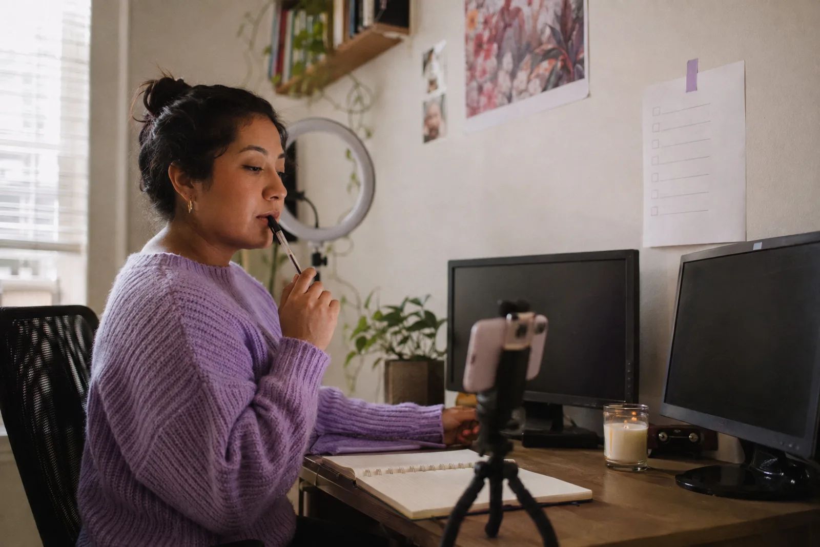 A creator at her desk with a pen at her lips, a blank checklist taped to the wall, two monitors and a phone on a tripod, thinking through the next deliverable.