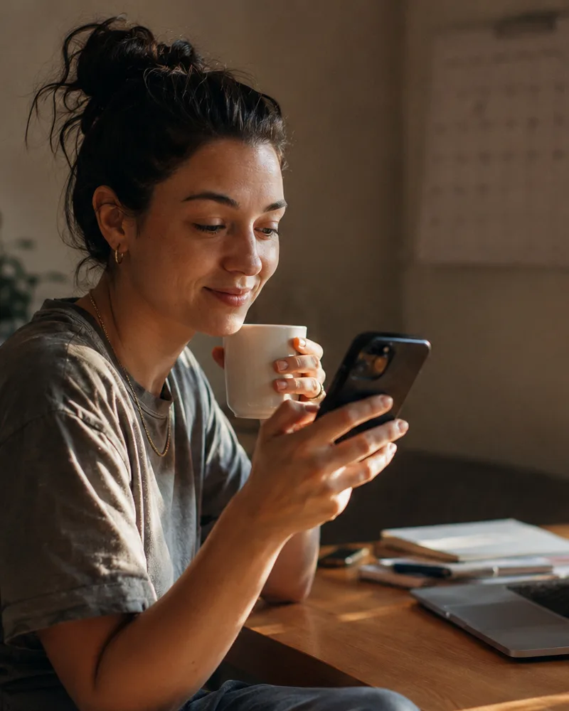 A creator at a sunlit desk with a coffee, glancing at a reminder on her phone with a small smile, laptop and planner beside her.