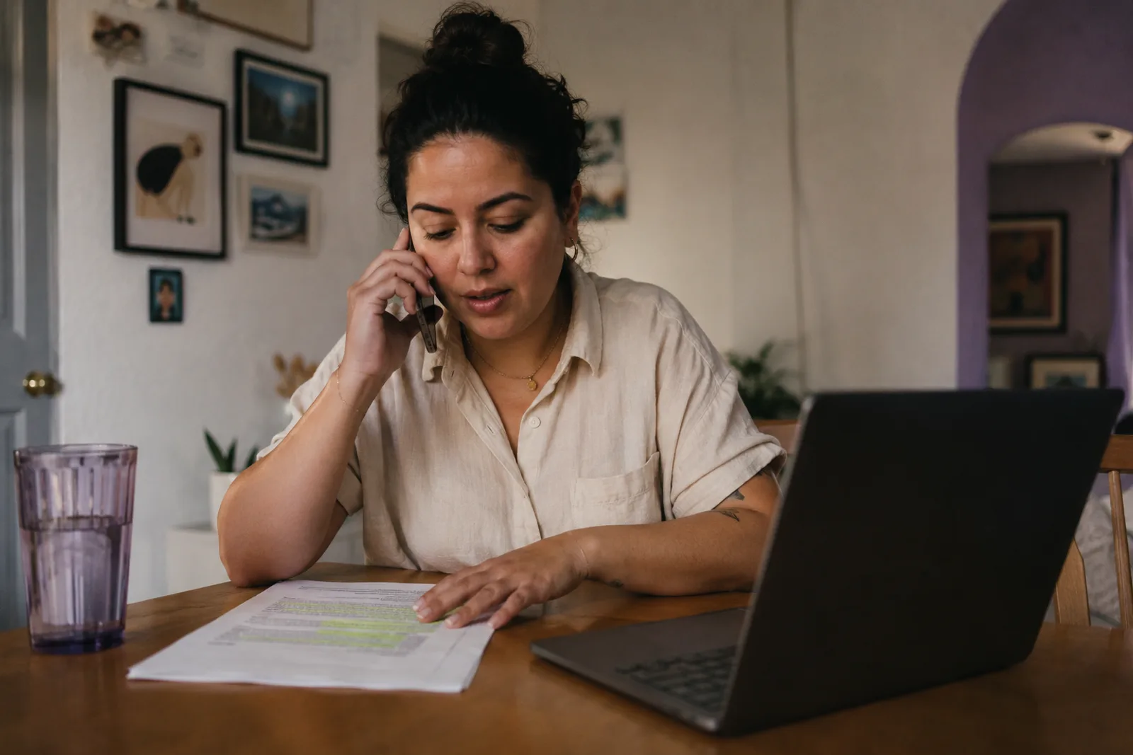 A creator at her dining table on a phone call, one hand on a printed page she has marked with a highlighter, laptop open beside her.