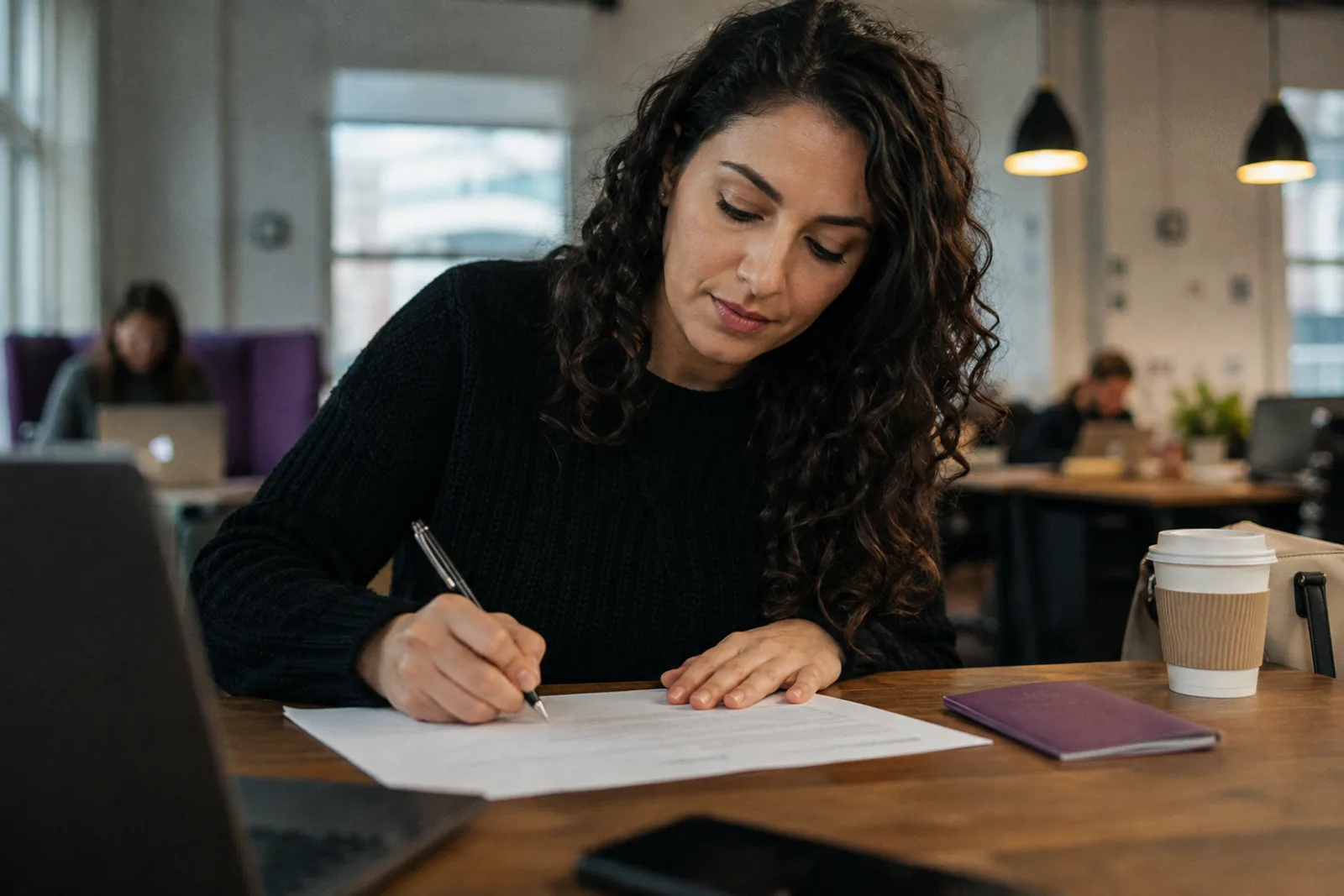 A creator at a co-working table signing paperwork with a pen, a folder and a coffee beside her, calm and unhurried.