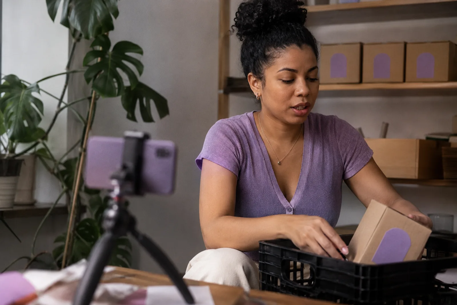 A creator in a small studio packing labeled product into a crate, three matching boxes lined up on the shelf behind her, a phone on a tripod in the foreground.