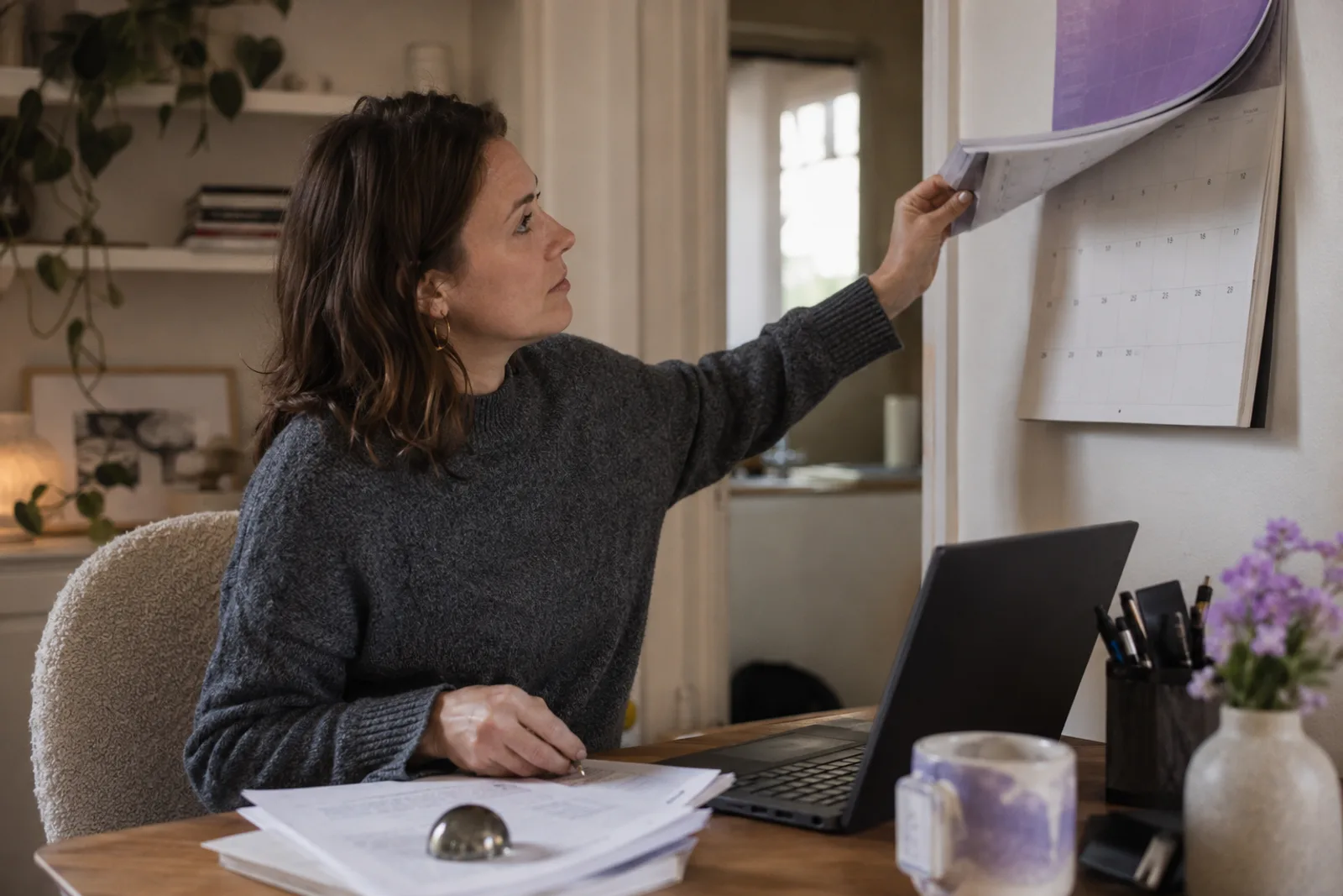 A creator in a home office flipping a wall calendar forward, laptop open, a small stack of signed pages under a glass paperweight.