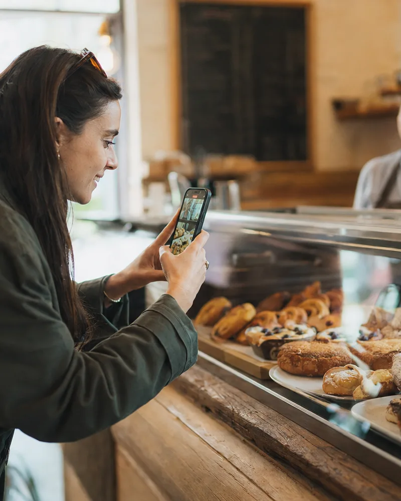 A creator leaning toward a bakery's pastry case, filming it on her phone, while the owner smiles from behind the counter.