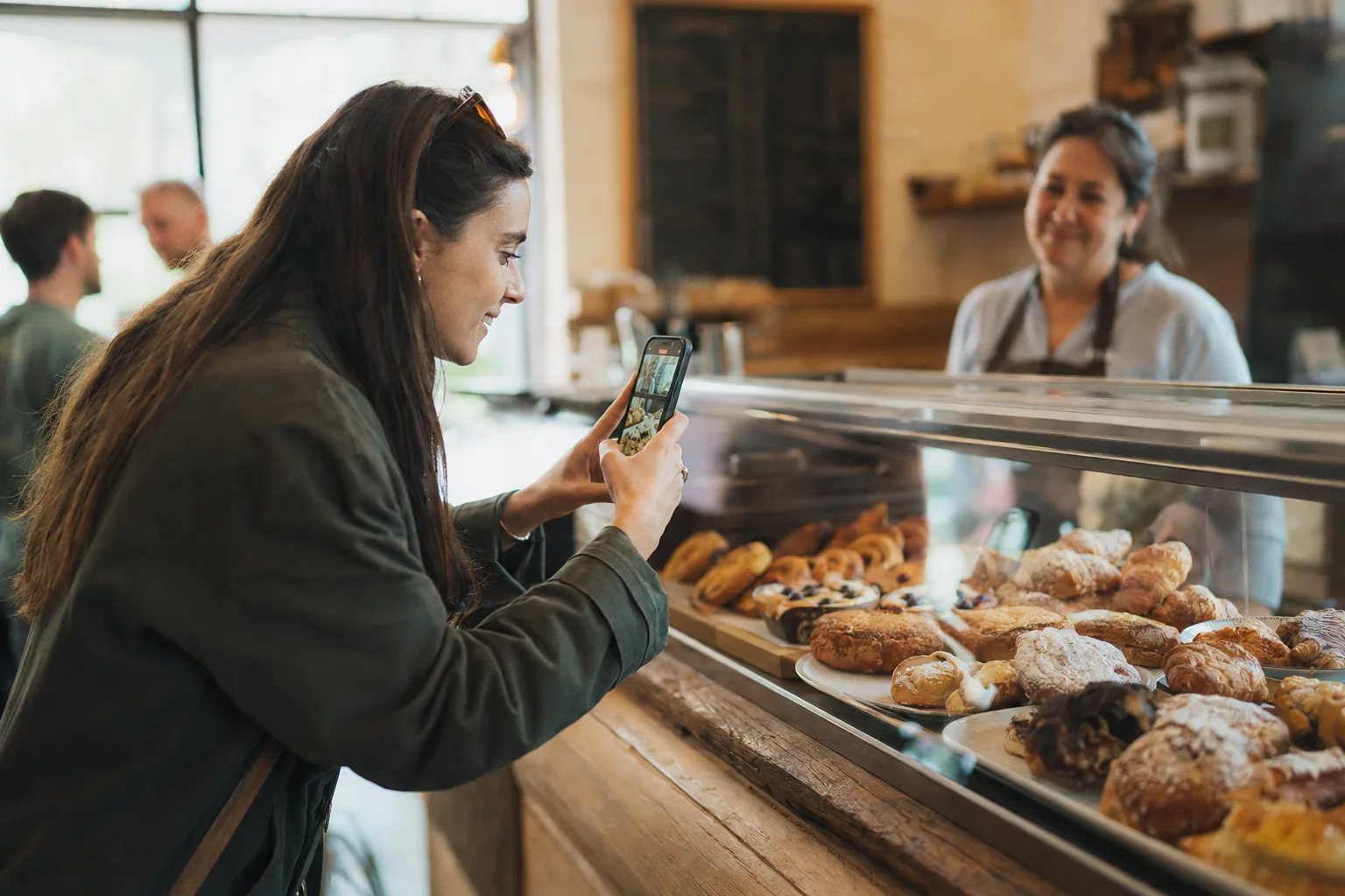 A creator leaning toward a bakery's pastry case, filming it on her phone, while the owner smiles from behind the counter.