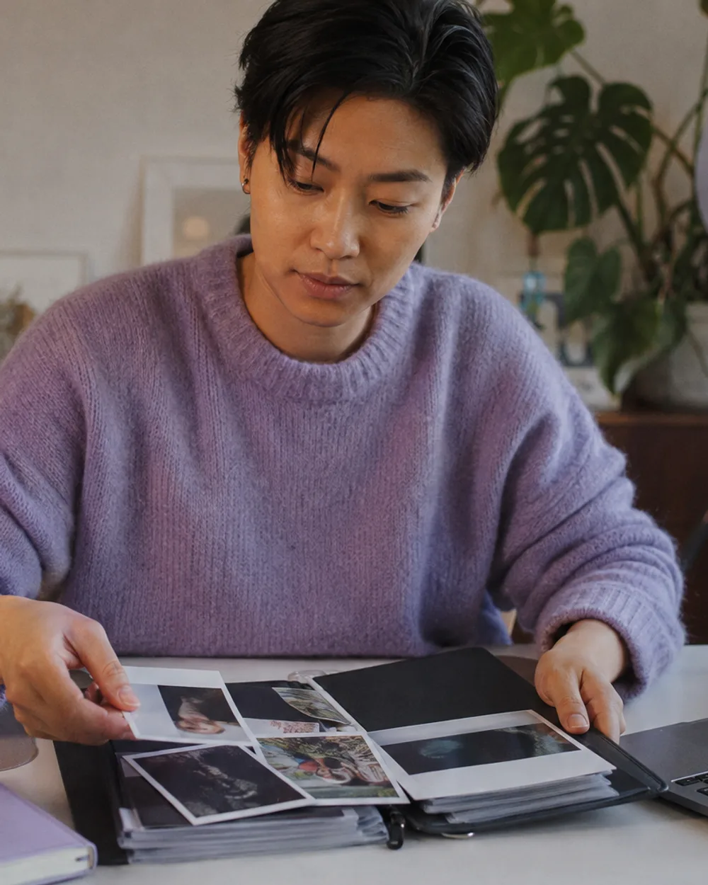A creator at a tidy desk arranging printed photos into a portfolio folder next to a laptop, a plant and a ring light behind them.