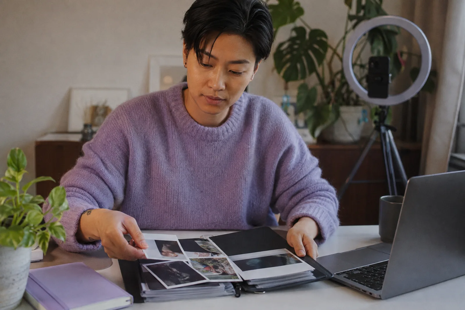 A creator at a tidy desk arranging printed photos into a portfolio folder next to a laptop, a plant and a ring light behind them.