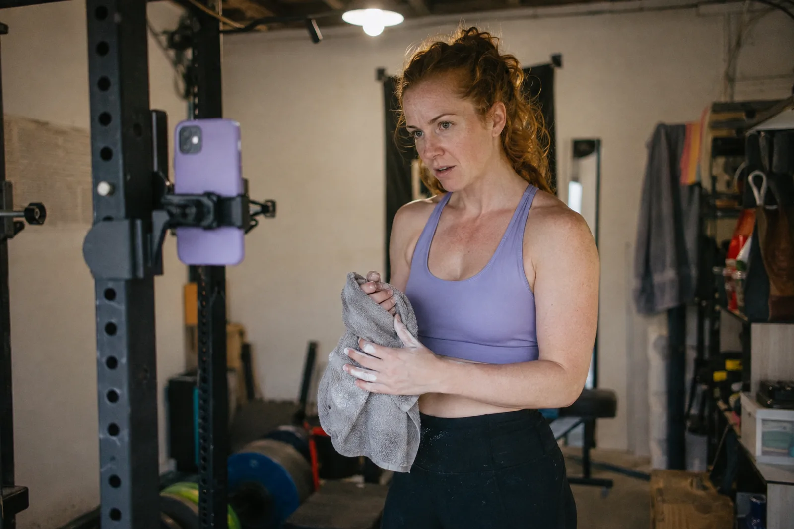 A creator in a garage gym wiping chalk from her hands with a towel while checking the shot on a phone clipped to the squat rack.