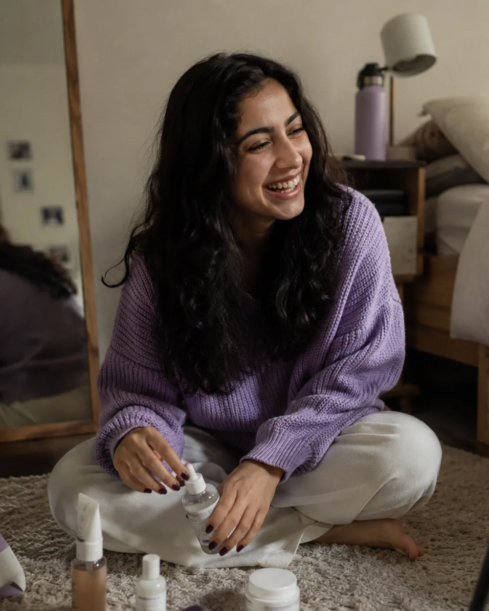 A young creator laughing on her bedroom floor, a phone on a small tripod in front of her and a few skincare bottles and one small package beside her.