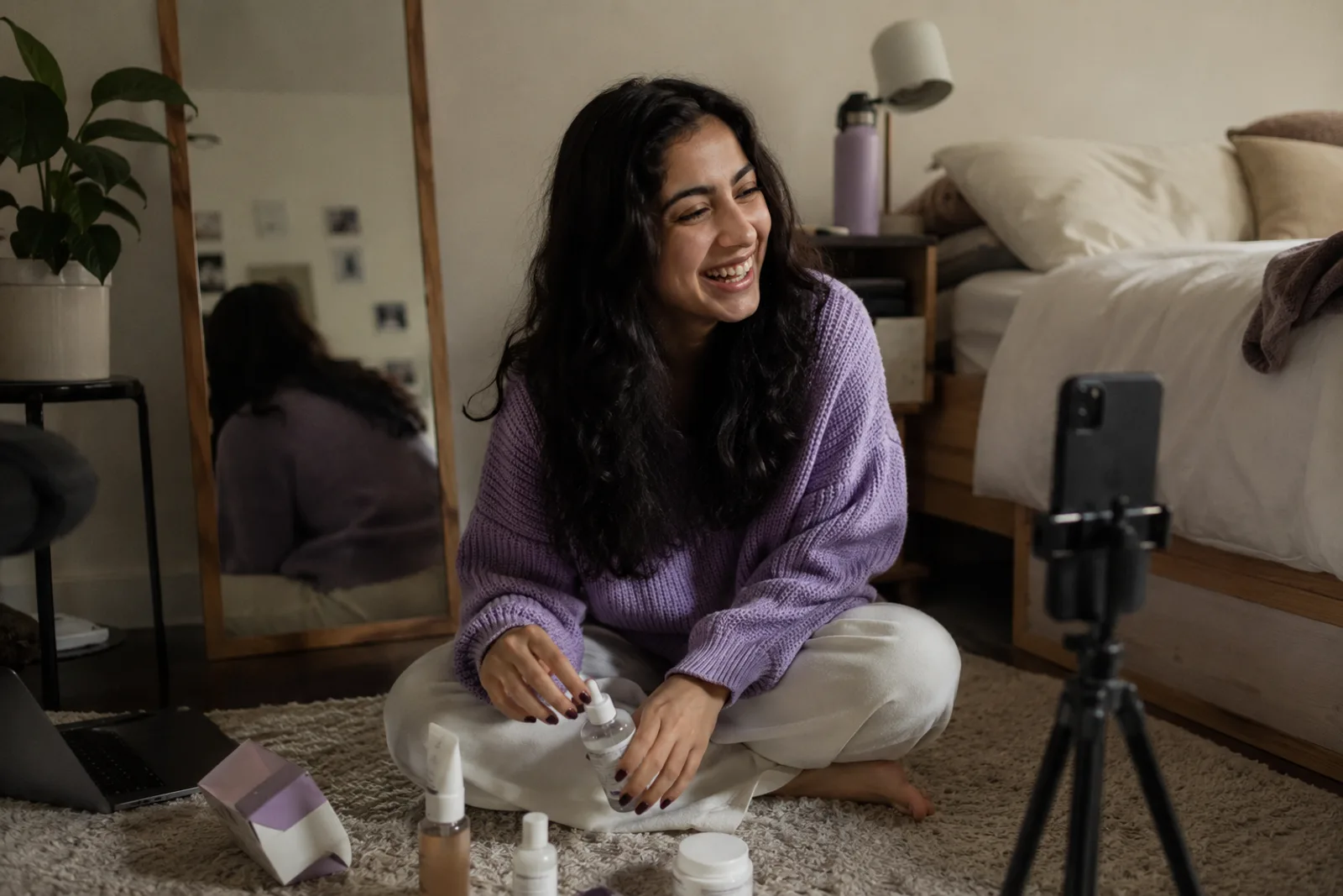 A young creator laughing on her bedroom floor, a phone on a small tripod in front of her and a few skincare bottles and one small package beside her.